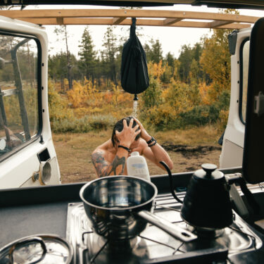 View from inside camper van on young hipster man take shower outdoors from portable shower.