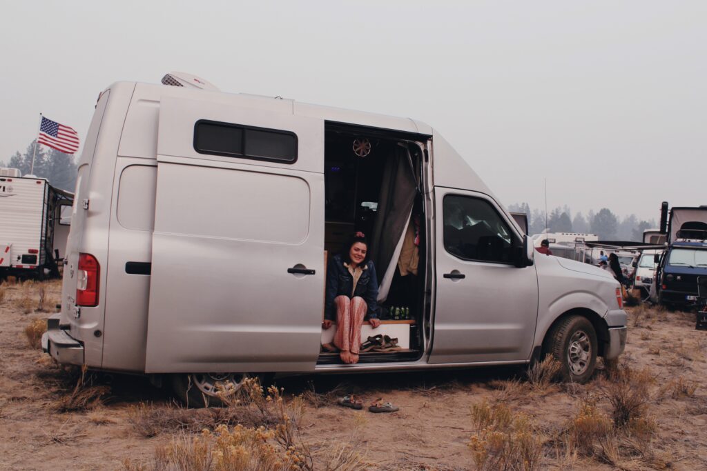 Exterior view of a young woman at a van life meetup, sitting in a DIY van build parked in the desert with several other DIY vans in the background.