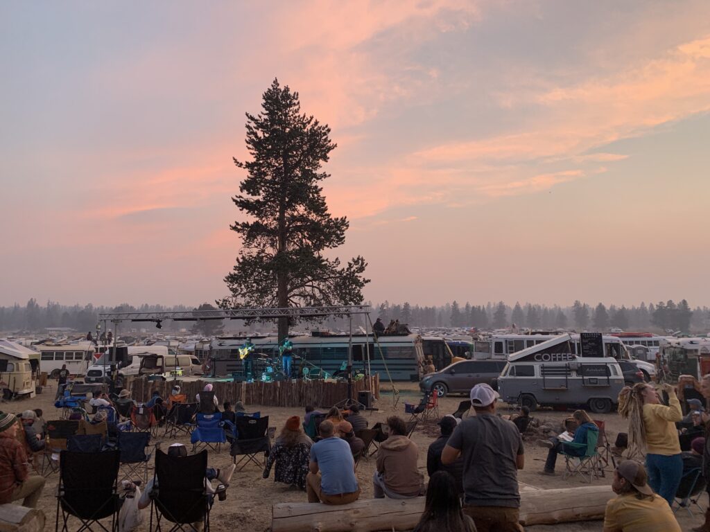 People gathered and sitting in front of a stage at a van life meetup as the sun sets with vans stretching back for a mile.