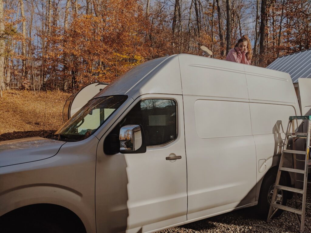 A girl perched on top of her Nissan NV 2500 van. Fall colors in the background.