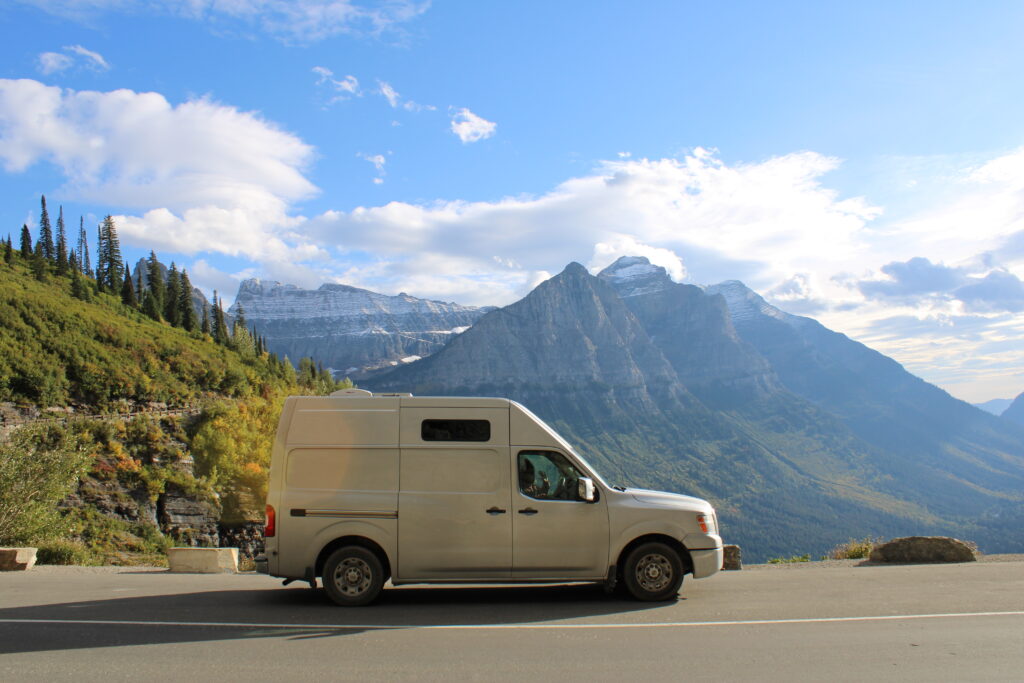 Nissan NV 2500 silver van on the side of the Going to the Sun Road in Glacier National Park. Snowy peaks are in the background.