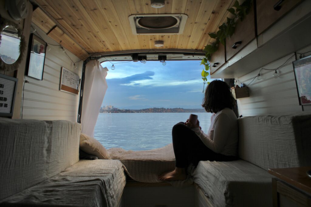 A girl sits in the back of her van with the back doors open, looking out at a lake view with a city across the way.