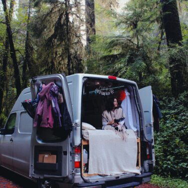 A camper van with the back doors open. A girl sits at the edge of the bed looking out the back with trees all around.