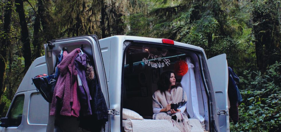 A camper van with the back doors open. A girl sits at the edge of the bed looking out the back with trees all around.