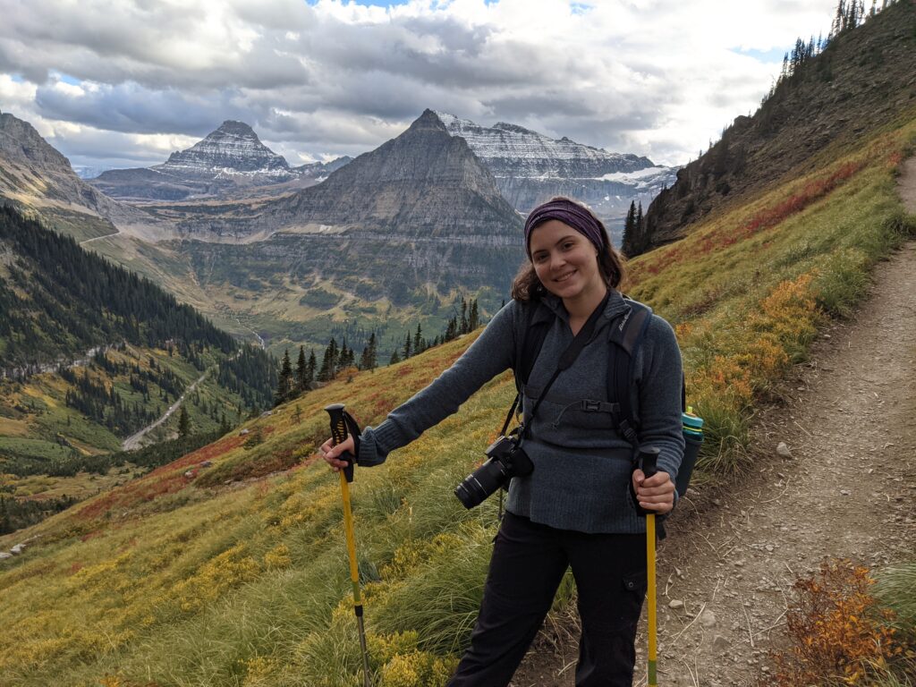 A girl with hiking gear poses on a trail in Glacier National Park with snowy peaks and mountain ranges in the background.