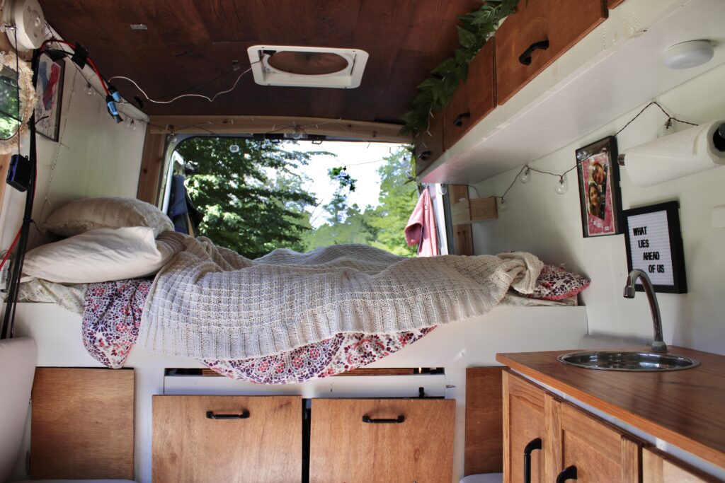 A stationary platform bed at the back of a camper van with the back doors open to trees. The bed had pink and white blankets on it and the van is decorated with lights and pictures with a white and wood theme.