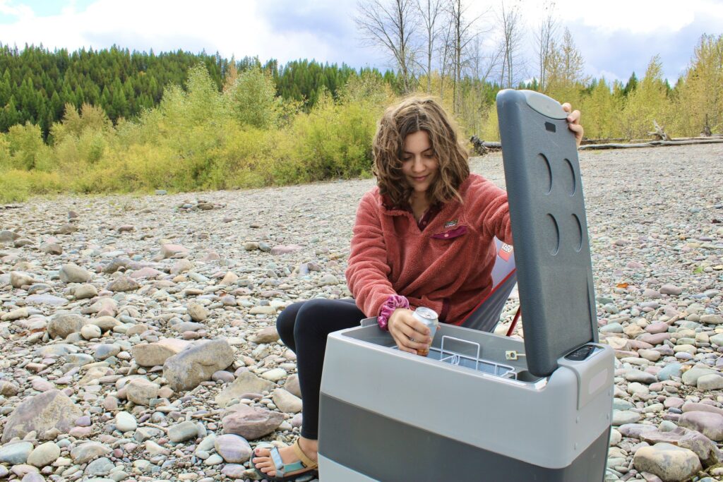 A girl sits by a rocky riverside in Montana, opening up a portable, chest-like fridge and pulling out a canned drink.