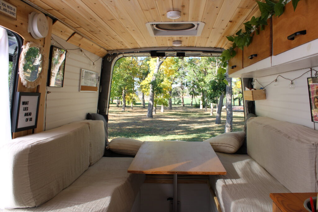 Interior of a camper van with bench seating on either wall and a wooden table in the middle. The van has a cedar plank ceiling and white walls, and the back doors are open to a park.