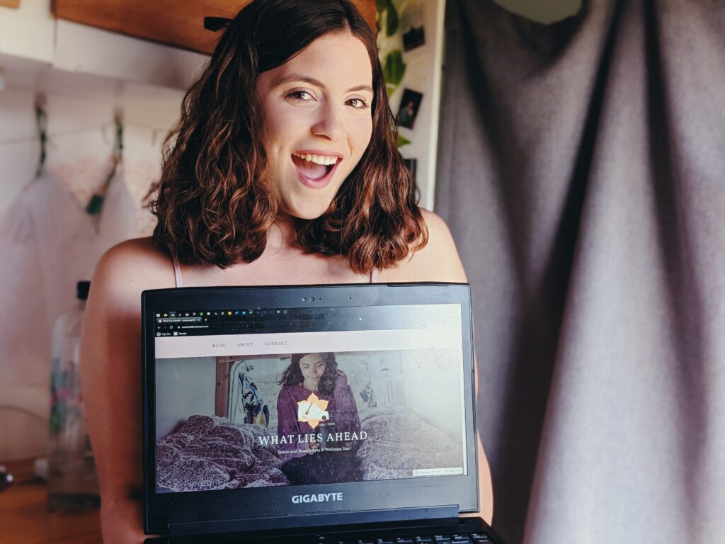A smiling girls standing in her camper van holds up a laptop with her website on it, entitled "What Lies Ahead."