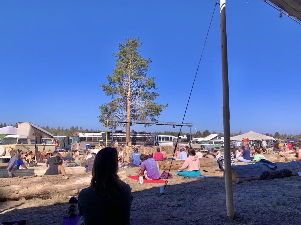 A group of people with yoga mats gathered outside for morning yoga at a van life meetup. There are camper vans and buses lined up behind the stage with the instructor.