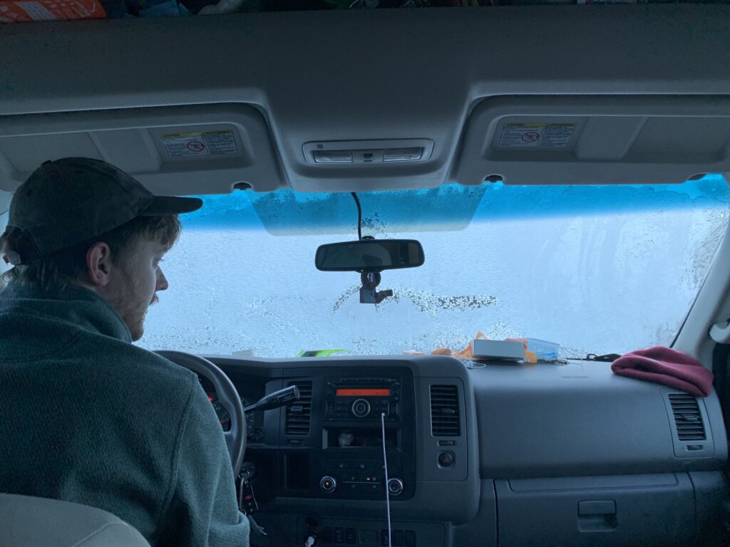A man sitting in the driver's seat of a camper van with a blanket of ice and frost covering the front windshield.