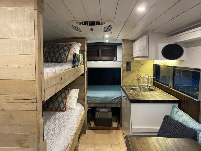 Interior of a camper van. There are two wooden bunk beds located along the left wall and one bunk bed above the full bed in the back. There is a kitchen to the right.