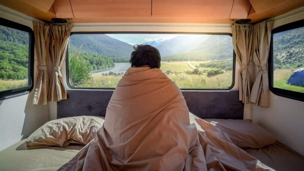 A person sitting inside their camper van bundled up in a blanket as they look out their window to a mountainous view.