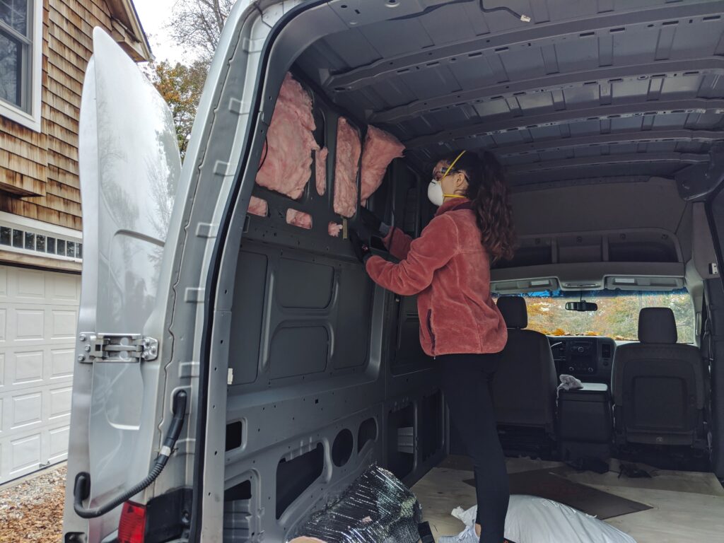 The interior of a bare camper van with a girl stuffing fiberglass insulation into the frame.