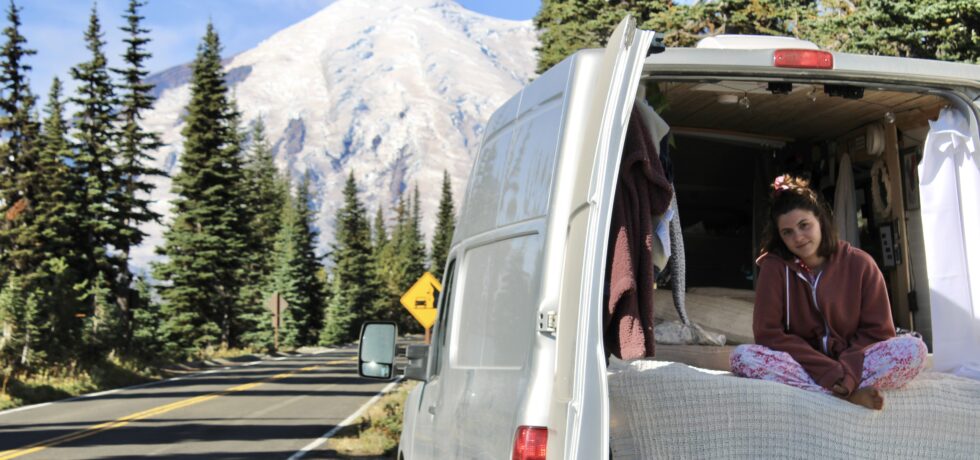 Woman sitting in the back of van conversion. The back door is open and there's a snow covered mountain in the background.