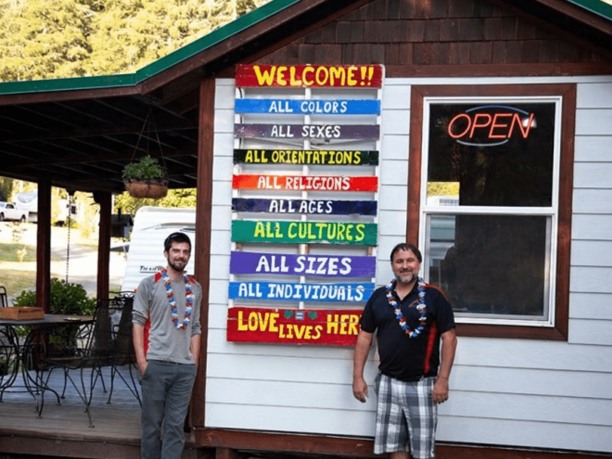 Two men stand outside of the campsite with a sign that reiterates that everyone is welcome. 