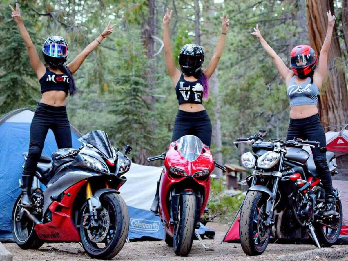 Three women throw their hands up as they stand on their motorcycles.
