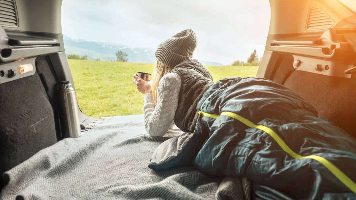 Girl resting in a sleeping bag in her van drinking out of a mug and looking out at mountains