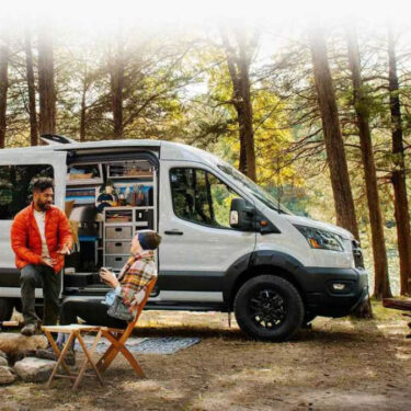 campers talking and relaxing outside of the 2023 Ford Transit Trail van set up in a wooded area by water