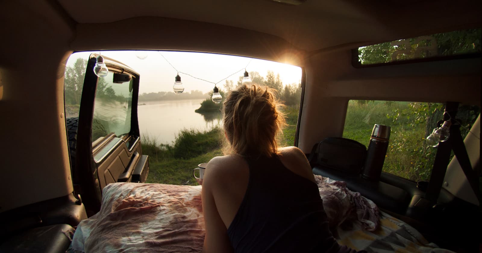 Woman laying in bed in van and drinking morning coffee. Beautiful calm lake in the background of this solo van life camping experience.