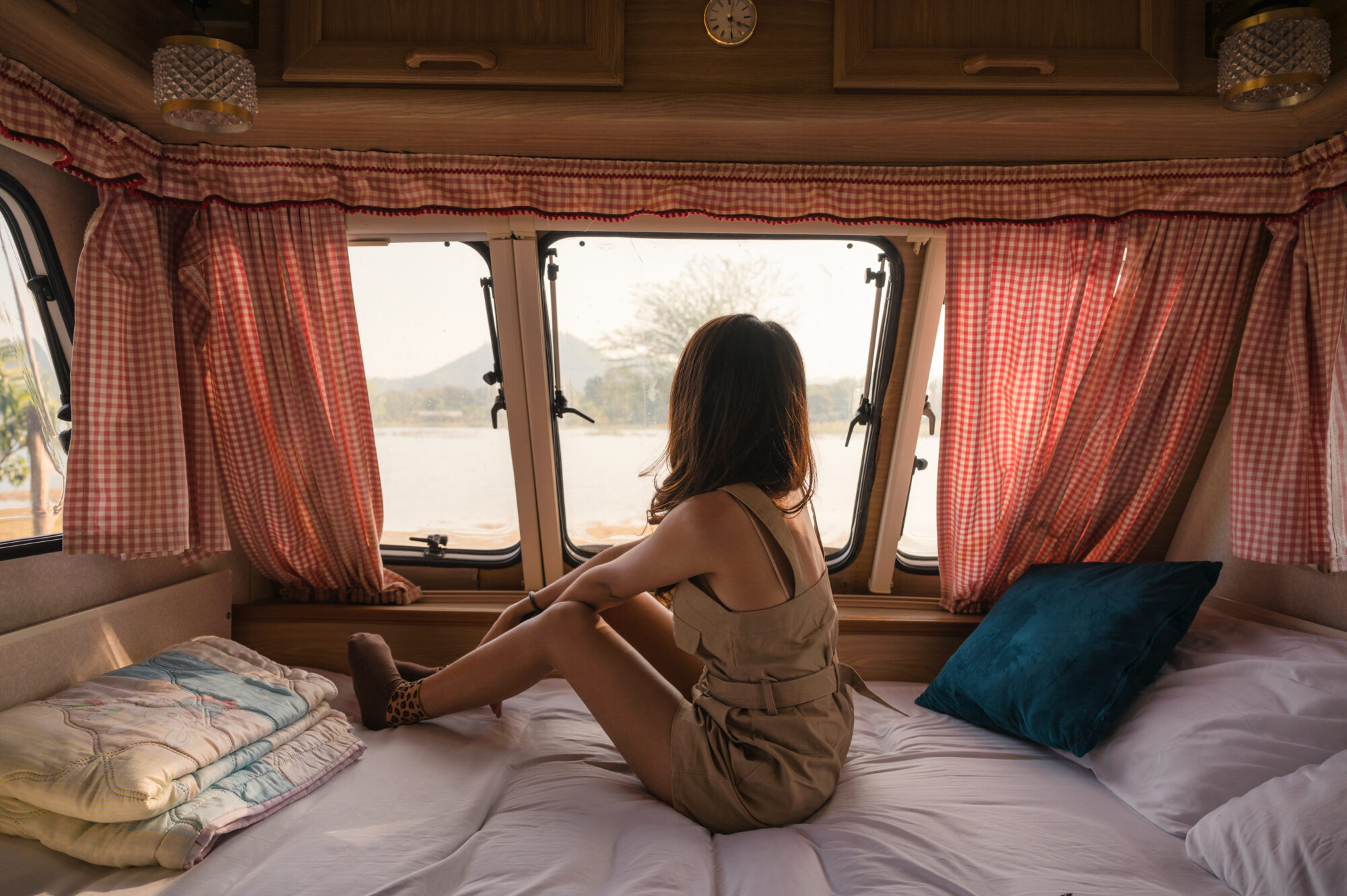  woman relaxing and looking at the view from her campervan bed. Checkered-pattern curtains are hanging as window covers.