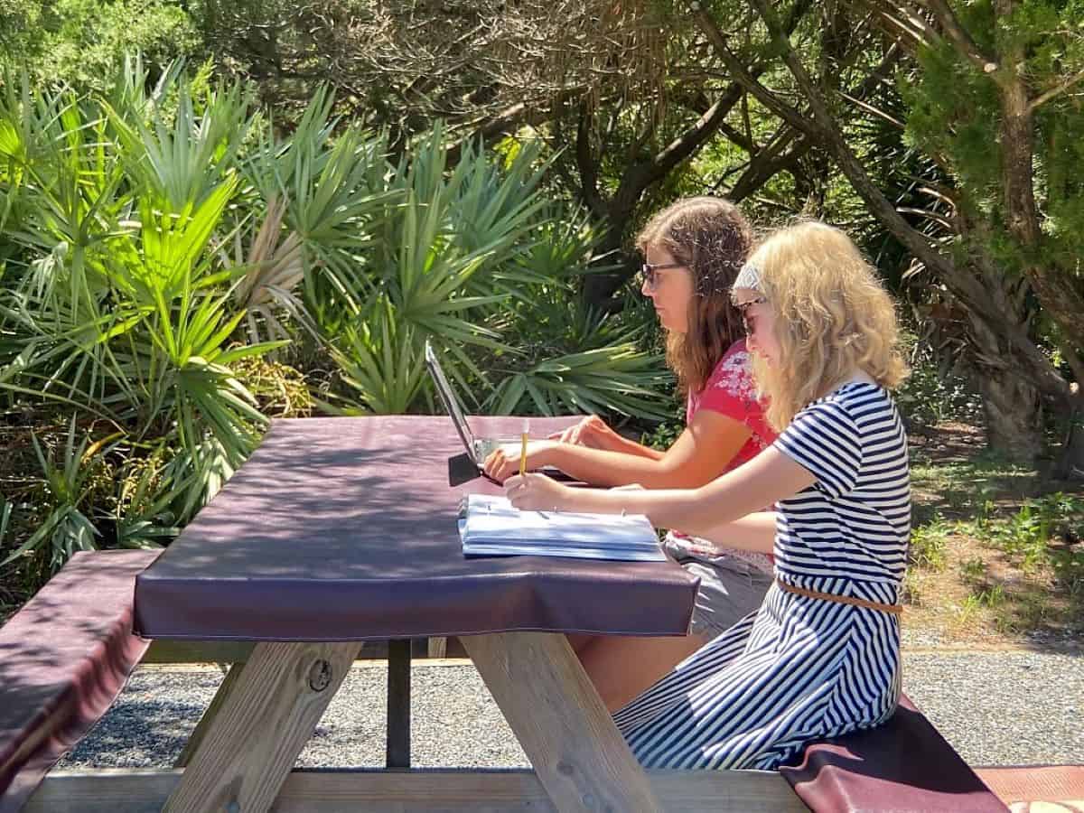 Woman working on the computer at a campsite picnic table while daughter is doing school.