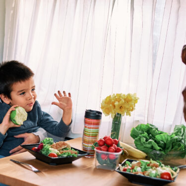 Multi-racial family eating salads-toddler son sitting on Mom's lap smiling at Dad across from the van dining table as he takes a picture of them. Communial areas like this make for ideal family-friendly van designs.