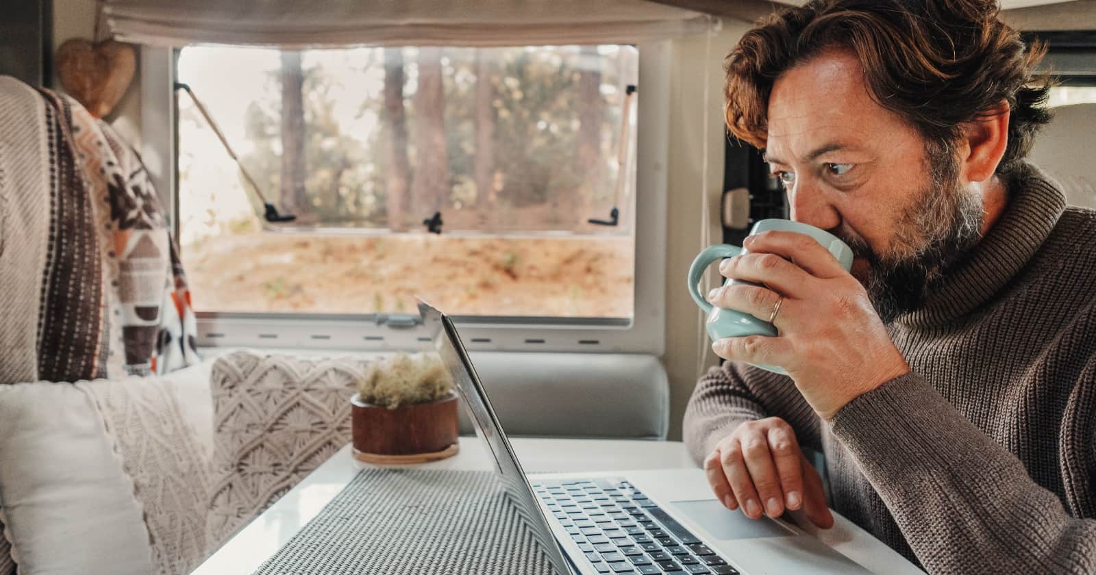 Mature man working on laptop computer inside a camper van with nature outdoors view outside the window. Van life offers the flexibility of a remote work schedule.