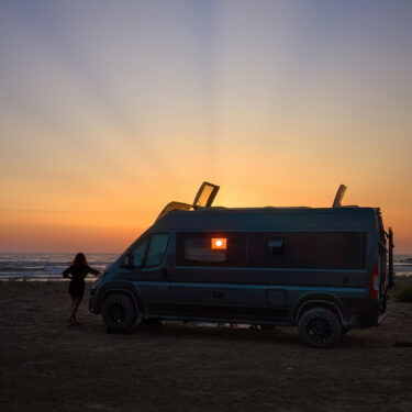 Woman standing outside a van on the beach at sunset.