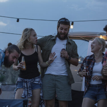 Group of laughing young men and women having drinks and chilling near trailer under garlands in campsite much like what attendees will experience at C.H.A.S.M. Fest