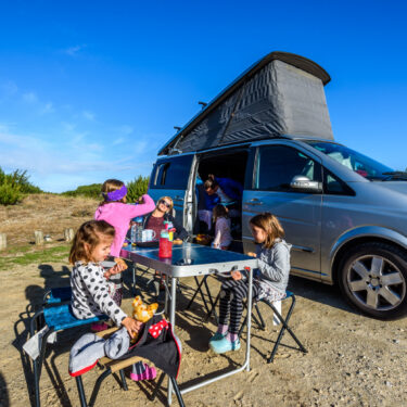 Family on vacation is sitting outsides of their camper van on camping chairs and table eating breakfast and other road trip snacks