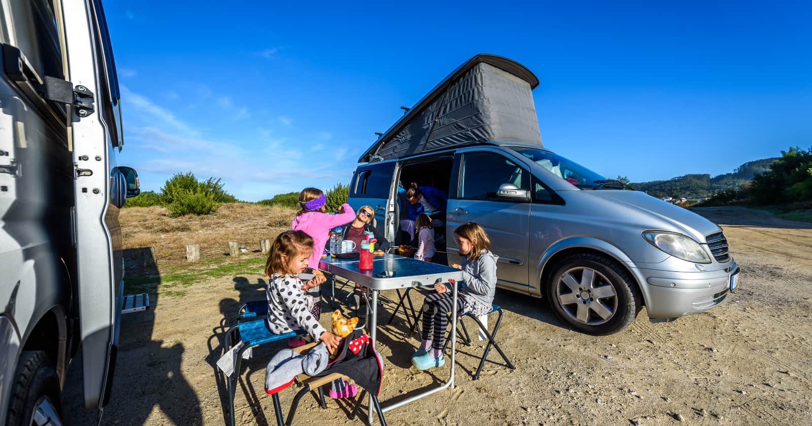 Family on vacation is sitting outsides of their camper van on camping chairs and table eating breakfast and other road trip snacks