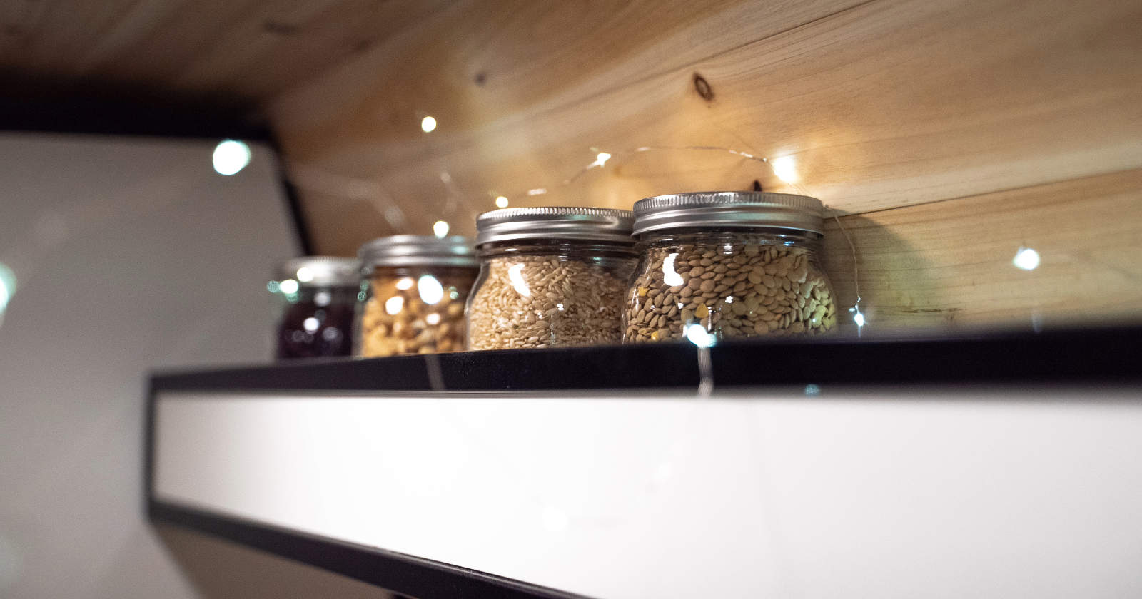 Glass jars filled with assorted grains and seeds, neatly lined on a black shelf against a wooden wall, with soft fairy lights reflecting in the background.