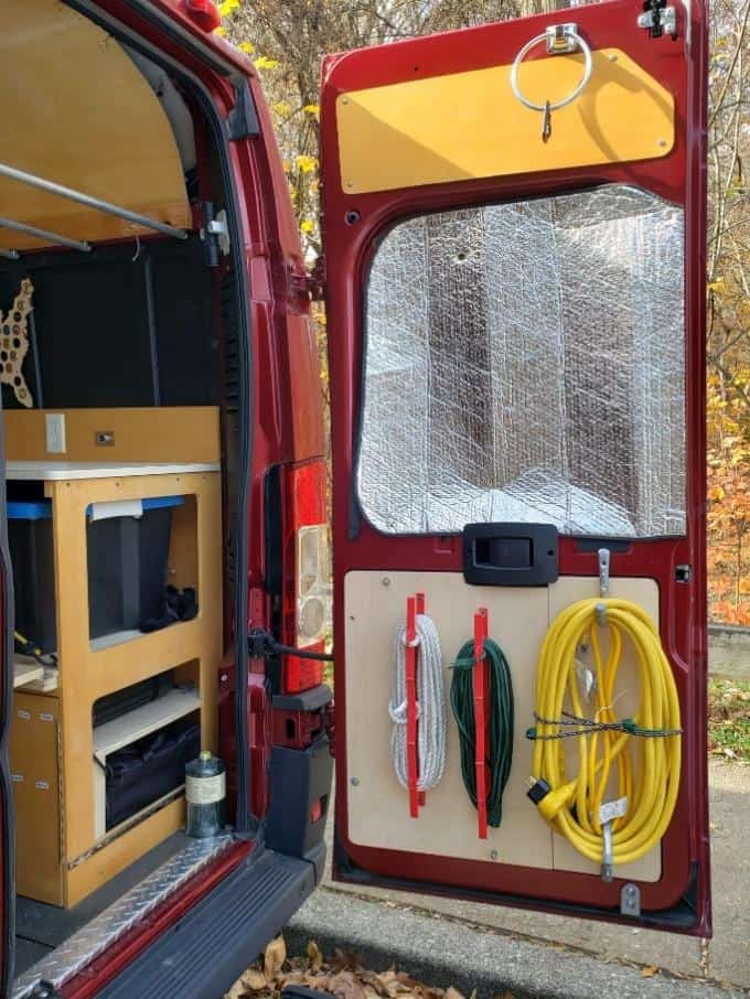 The rear view of a red van with its back door open, revealing a customized storage system. The left side features wooden cabinetry with compartments, including a small fridge and storage spaces. The right door is fitted with hooks holding a green hose, a red cable, and a yellow electrical cord neatly coiled. Above the storage, a reflective silver sunshade is attached to the window for insulation, and a circular metal ring hangs from the top of the door. The van is parked outside with fallen leaves on the ground, indicating autumn.