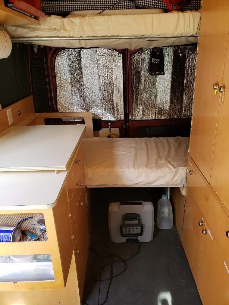 Interior view of a customized camper van. On the left, a wooden cabinet with multiple drawers and a small white countertop. The right side features a raised bed with a beige mattress, under which a portable heating unit and a large water jug are stored. The windows are covered with reflective insulation material, and a black hand towel hangs on a hook. A black curtain separates the sleeping area from the front of the van. The flooring is gray carpet, and there's a sense of efficient use of space.