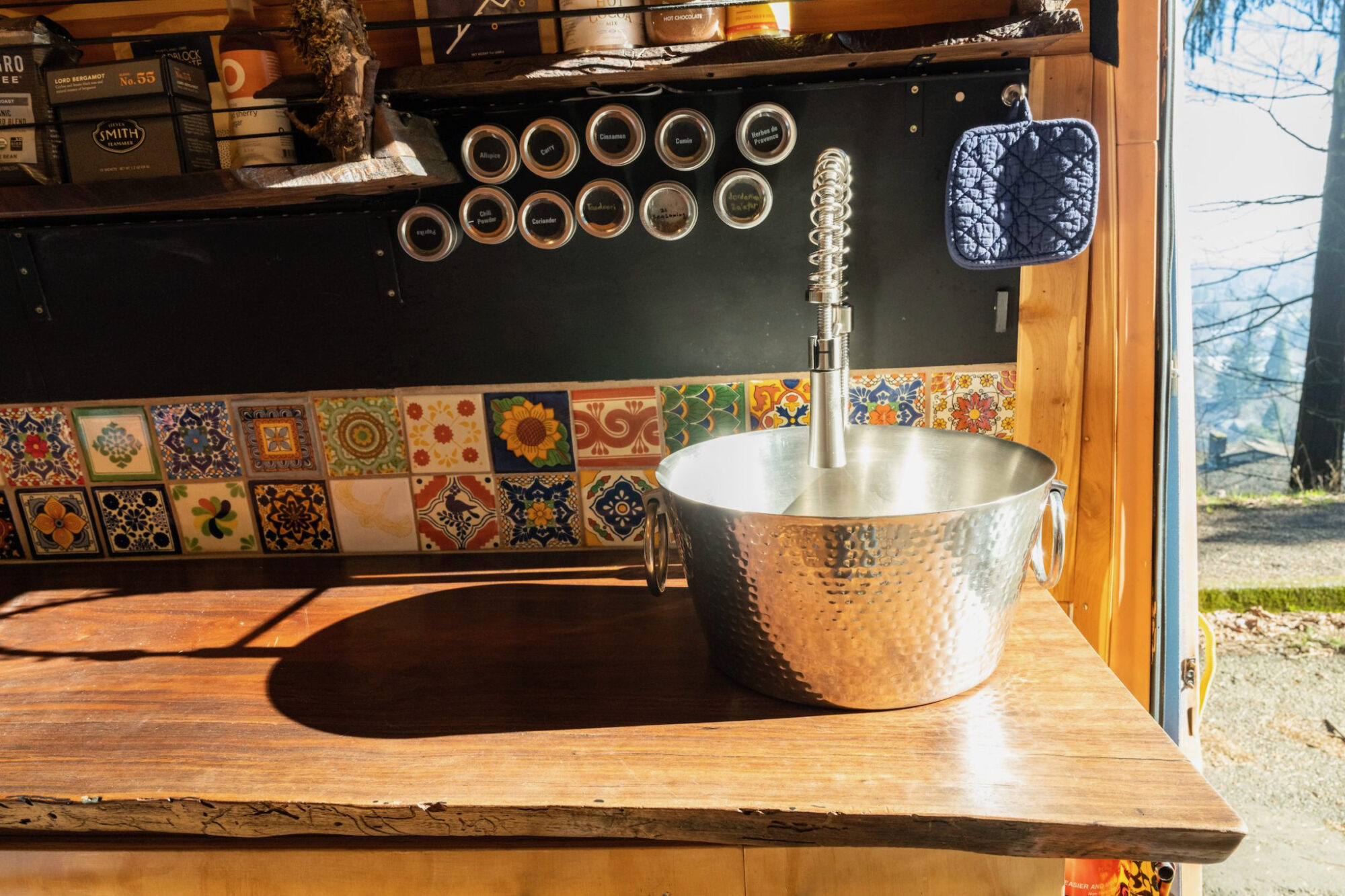 An eclectic kitchen corner featuring a hammered copper sink with a high-arc faucet, set against a backdrop of vibrant, multicolored Spanish tiles. The countertop is a slab of live-edge walnut, its natural edges and deep wood grain illuminated by the streaming sunlight from the open door. 
