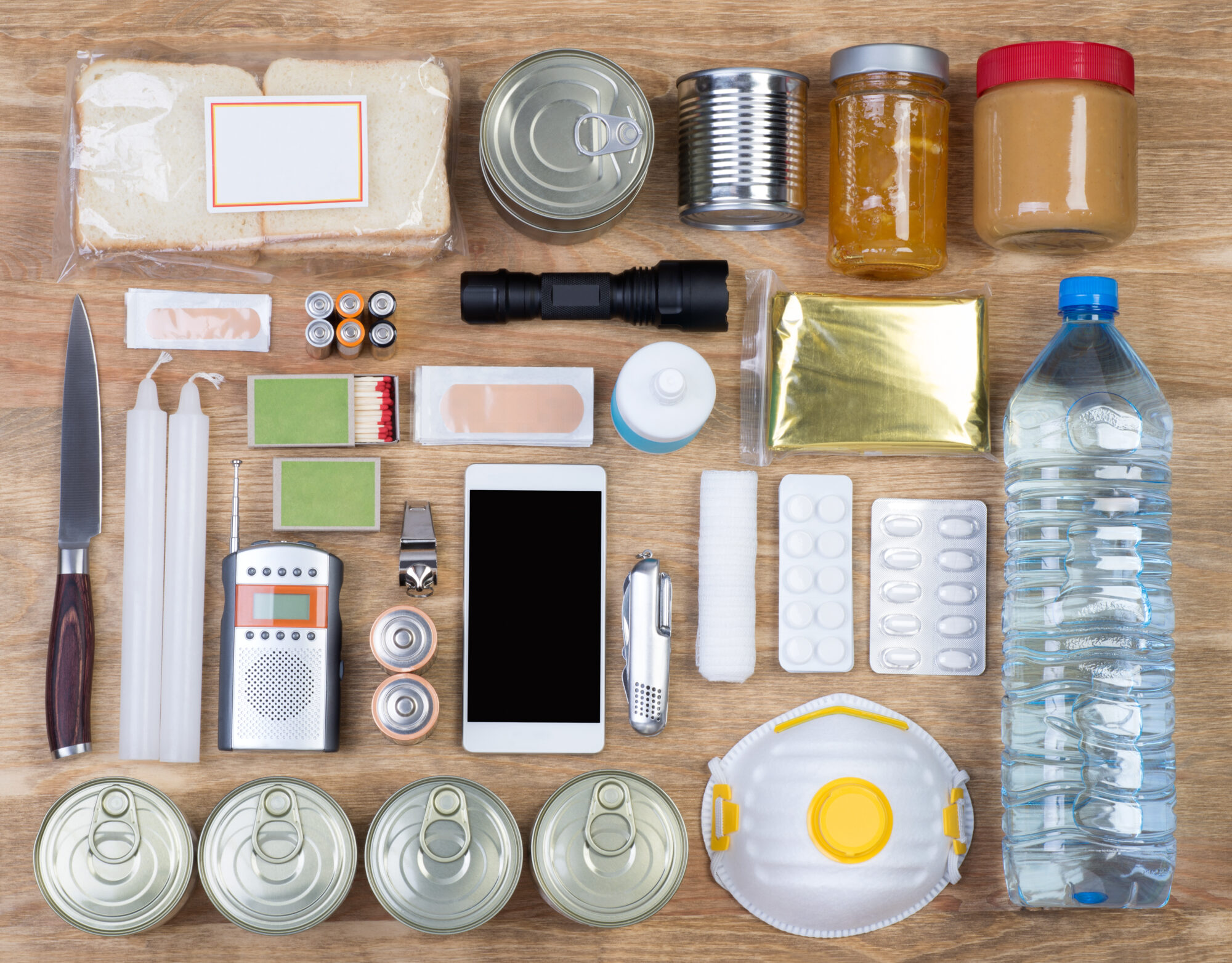A well-organized survival kit displayed on a wooden surface. The kit includes essential items such as a can opener, canned food, jars of peanut butter and jam, a flashlight, batteries, candles, matches, green scrubbing pads, soap, a pocket knife, a radio, a whistle, a smartphone, adhesive bandages, tablets in blister packs, a roll of gauze, a dust mask, and a large bottle of water.
